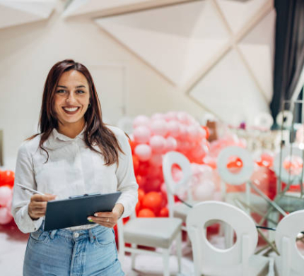 Portrait of cheerful and beautiful female party organizer standing in restaurant surrounded by balloon decoration and holding clipboard